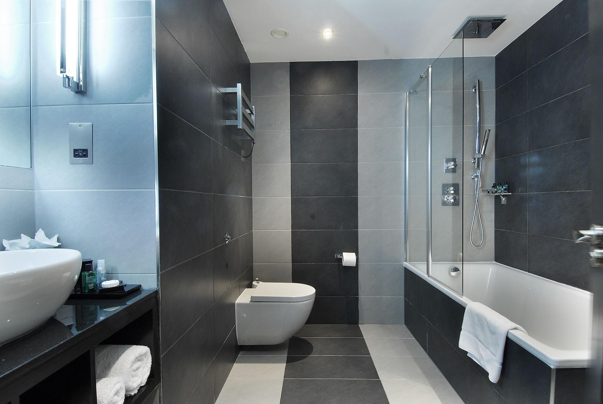 ホテル Montcalm Brewery Townhouse ロンドン エクステリア 写真 The photo shows a modern bathroom featuring a sleek design. The walls are tiled in a combination of dark gray and light gray, creating a contemporary look. On the left, there is a countertop with a basin and some toiletries placed on it. The middle s