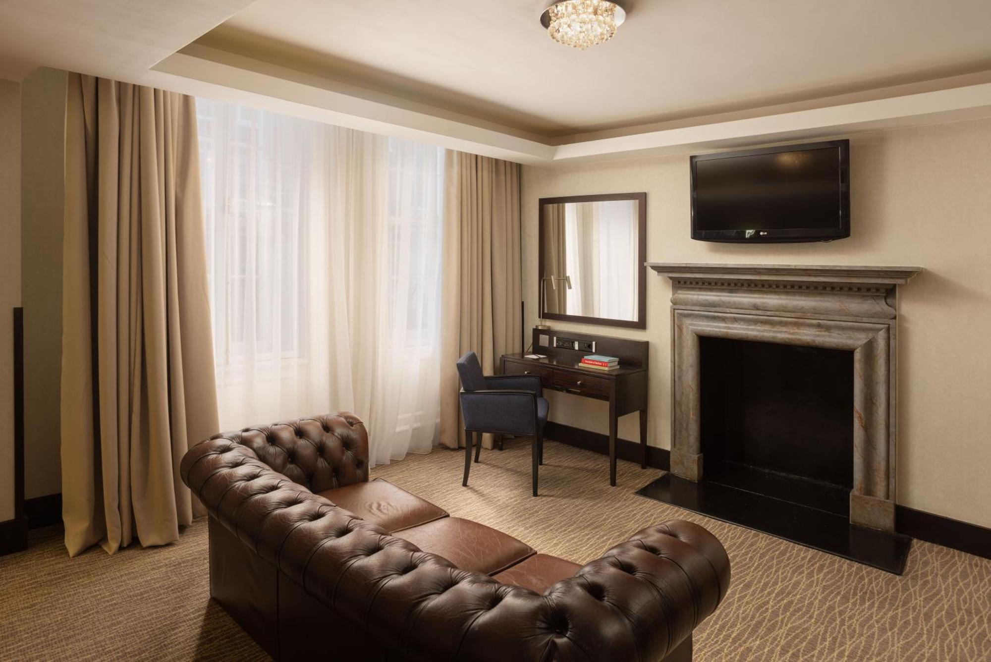 ホテル Montcalm Brewery Townhouse ロンドン エクステリア 写真 The photo shows a stylish interior of a room, likely a living area or a hotel suite. It features a dark leather chesterfield sofa with tufted upholstery. Opposite the sofa is a small wooden desk with a chair, positioned near a wall-mounted flat-scree