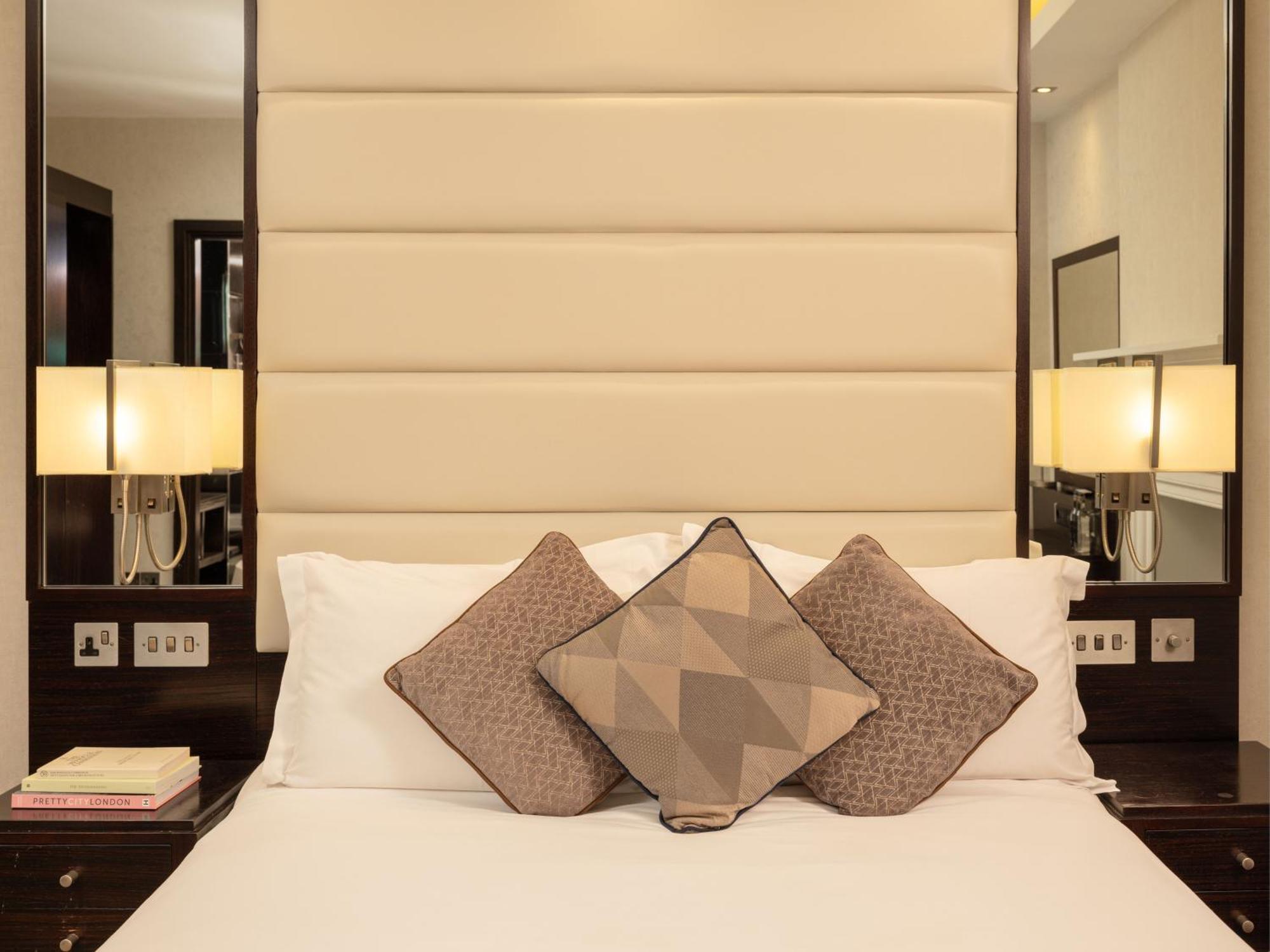 ホテル Montcalm Brewery Townhouse ロンドン エクステリア 写真 The image shows a cozy bedroom setting. The bed features a headboard upholstered in a light beige fabric. It is adorned with three decorative pillows in varying patterns and colors, placed on white bedding. On either side of the bed, there are modern