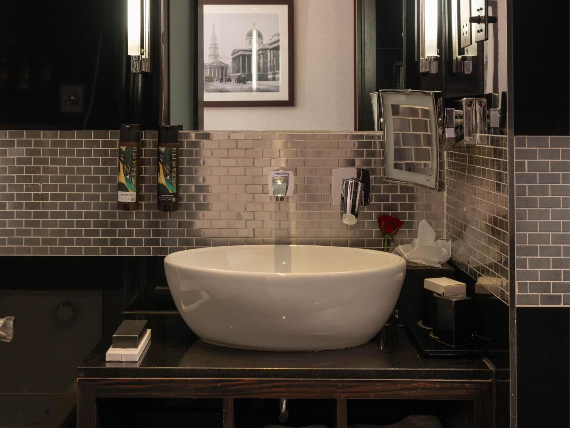 ホテル Montcalm Brewery Townhouse ロンドン エクステリア 写真 The photo shows a modern bathroom with a stylish design. It features a large, round white sink placed on a dark wooden vanity. The walls are adorned with a mix of gray and silver tile, creating a sleek and contemporary look. Above the sink, there is 
