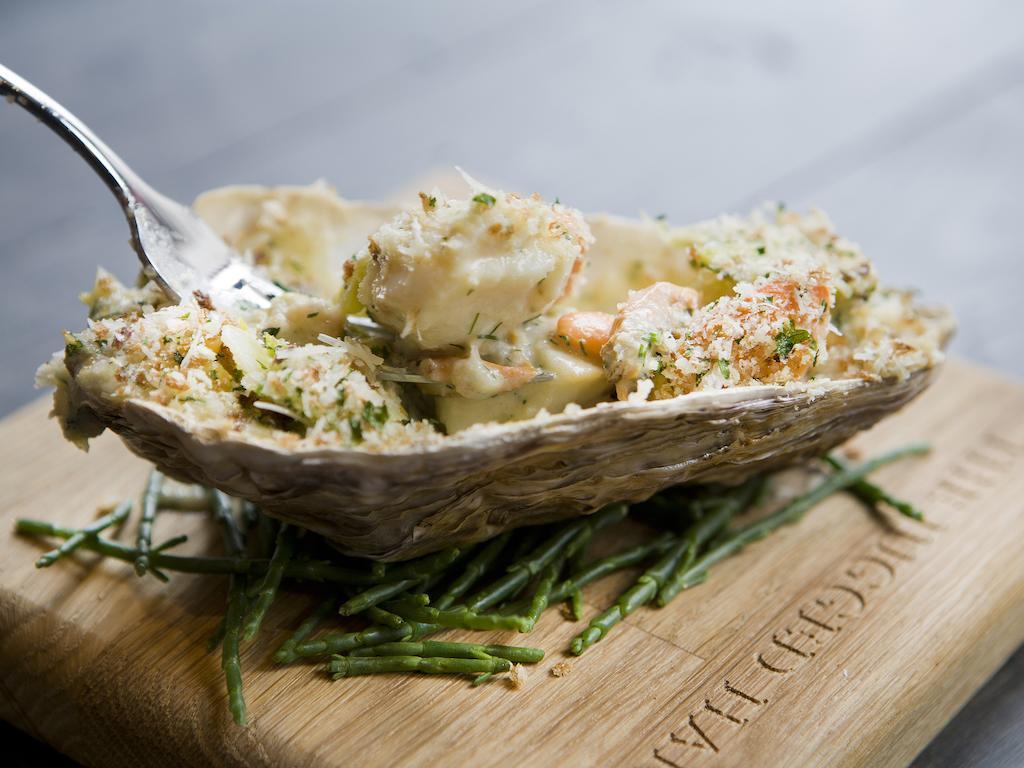 ホテル Montcalm Brewery Townhouse ロンドン エクステリア 写真 The photo shows a large oyster shell filled with a mixture that appears to include seafood, herbs, and breadcrumbs. The dish is presented on a wooden board with some greenery, possibly a type of seaweed or garnishing. A fork is placed beside the oyst