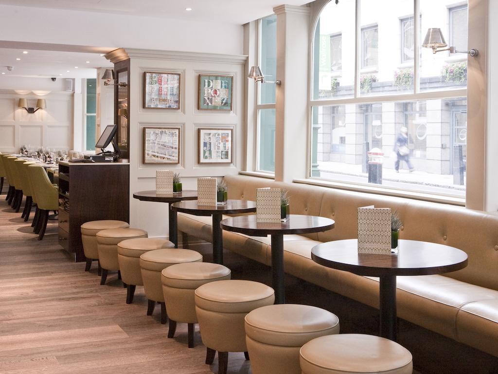 ホテル Montcalm Brewery Townhouse ロンドン エクステリア 写真 The photo displays a modern café or restaurant interior. There is a cozy seating area featuring light-colored, upholstered benches along the wall, accompanied by small round tables. The tables are paired with circular stools. The decor includes frame